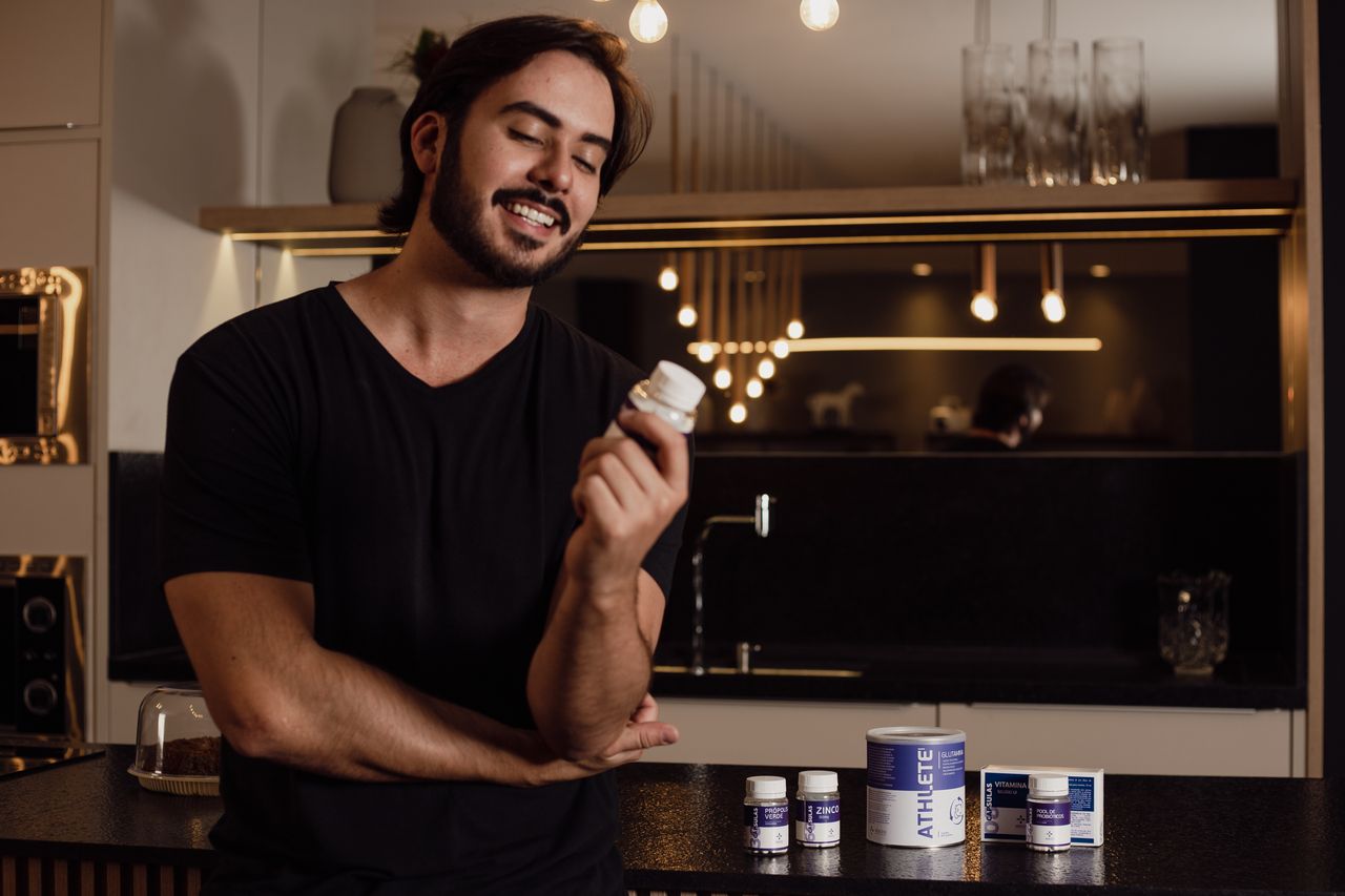 A smiling man in a kitchen holds a bottle of supplements, showcasing health and wellness.