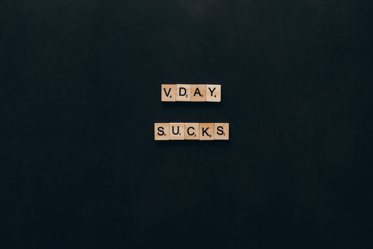 Scrabble tiles spell out 'V. Day Sucks' on a black background, conveying an anti-Valentine's sentiment.